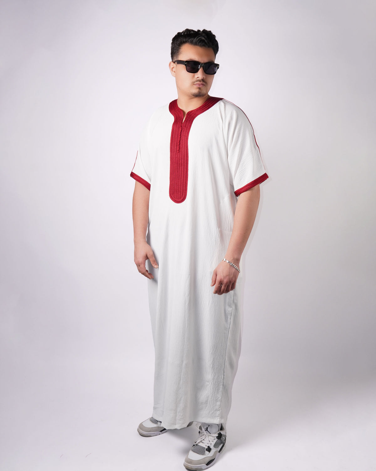 Man standing wearing a full-length white thobe with short sleeves and maroon embroidered trim.