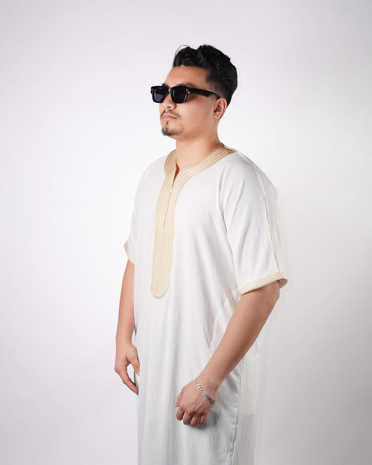 Man wearing sunglasses stands facing slightly away, modelling a white short-sleeve thobe with gold embroidery detail on the neckline and cuffs.