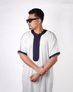 Man wearing sunglasses looking over his shoulder with arms crossed, modelling a white short-sleeve thobe with navy blue embroidered trim.