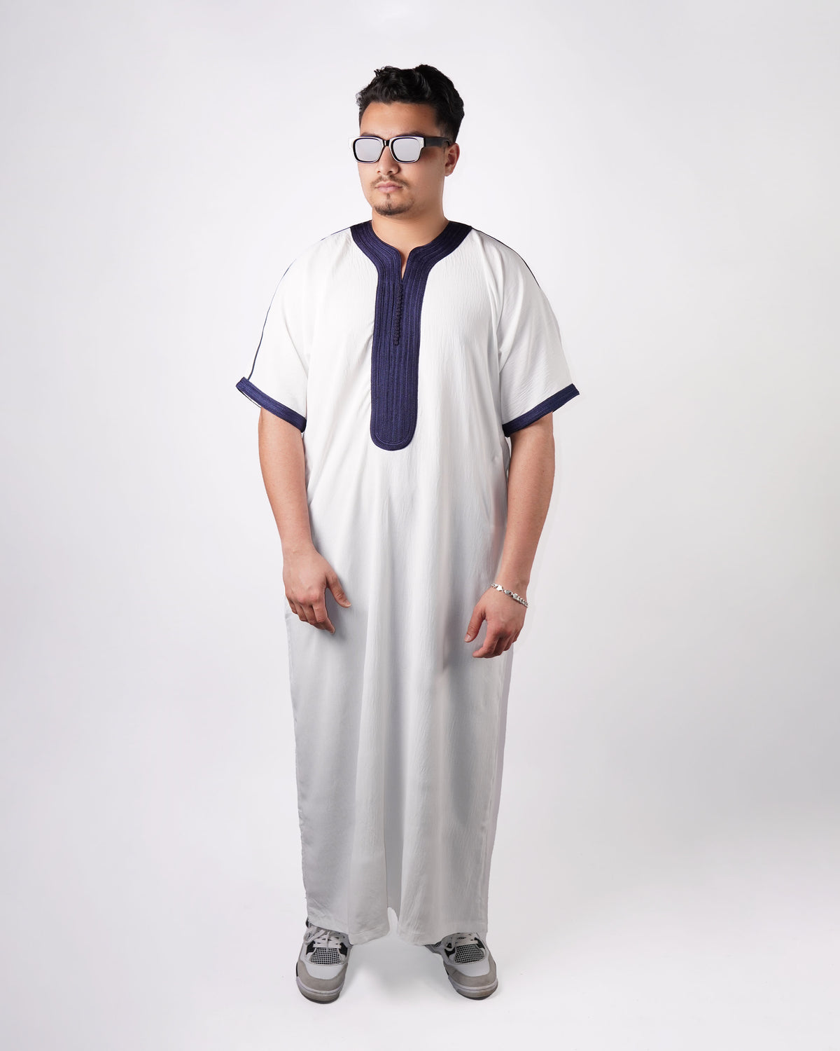 Full-length front view of a man wearing sunglasses and sneakers, modelling a white short-sleeve thobe with navy blue embroidered trim.