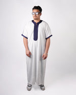 Full-length front view of a man wearing sunglasses and sneakers, modelling a white short-sleeve thobe with navy blue embroidered trim.