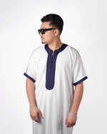 Man wearing sunglasses in side profile, modelling a white short-sleeve thobe featuring navy blue embroidered trim.