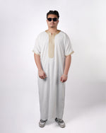 Full-length front view of a man wearing sunglasses modelling a white short-sleeve gandoura featuring gold embroidered trim, paired with sneakers.