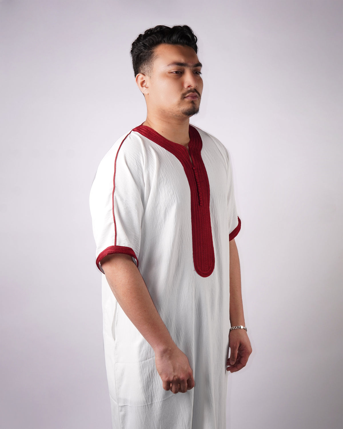 Man standing in side profile wearing a white short-sleeve thobe with maroon embroidered trim.