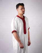 Man standing in side profile wearing a white short-sleeve thobe with maroon embroidered trim.