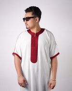 Man wearing sunglasses standing in a white short-sleeve gandoura with maroon embroidered trim, looking over his shoulder.