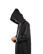 Side view of a man wearing a hooded black thobe with silver trim, hood pulled up.