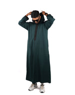 Man standing full length wearing a hooded green thobe with black trim, adjusting the hood.