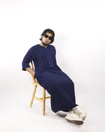 Man casually sitting in a stylish navy blue Moroccan thobe, showcasing comfortable Muslim male wear, paired with sunglasses and white sneakers.