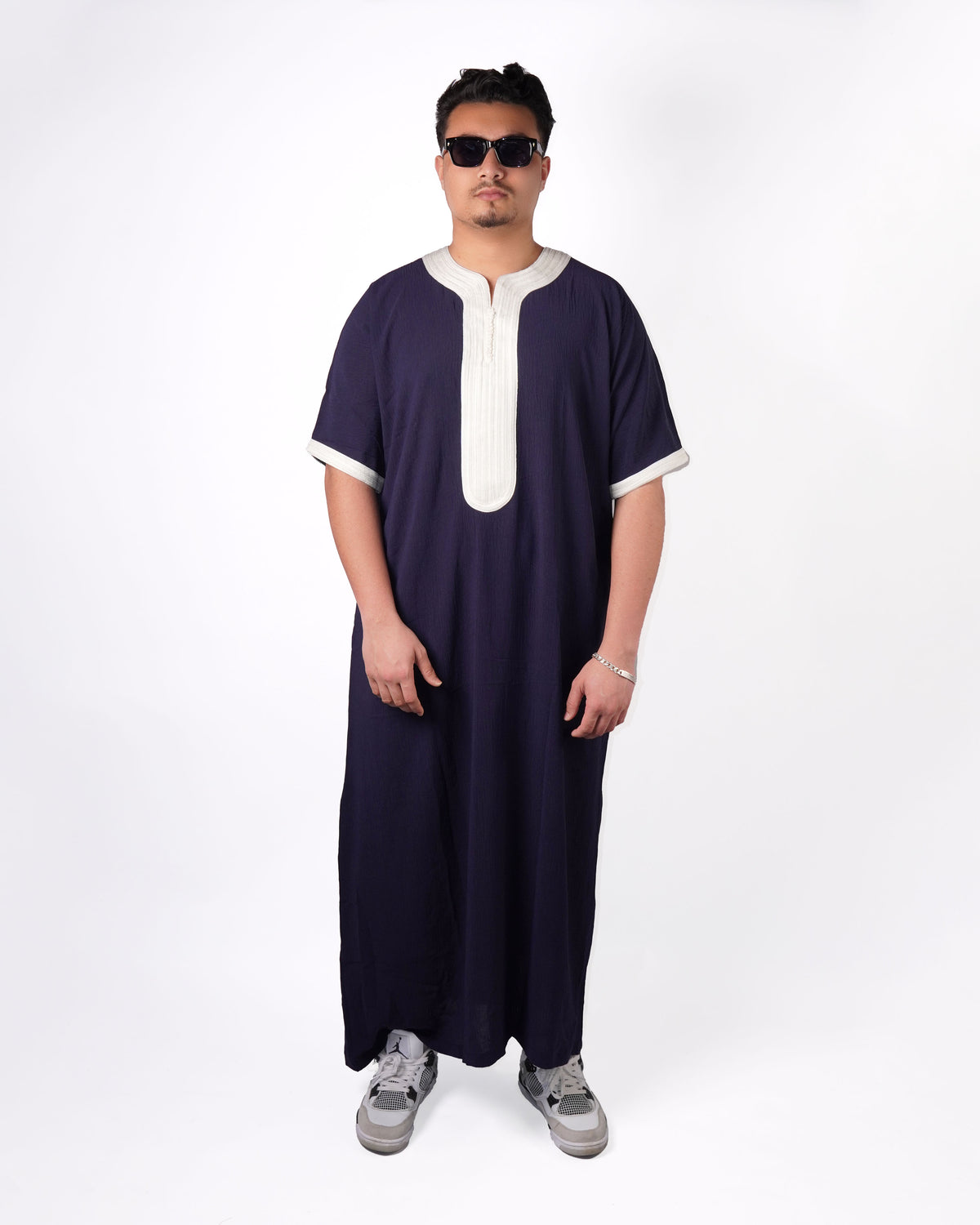 Full-length front view of a man wearing sunglasses and sneakers, modelling a navy blue short-sleeve thobe with light grey contrast trim.